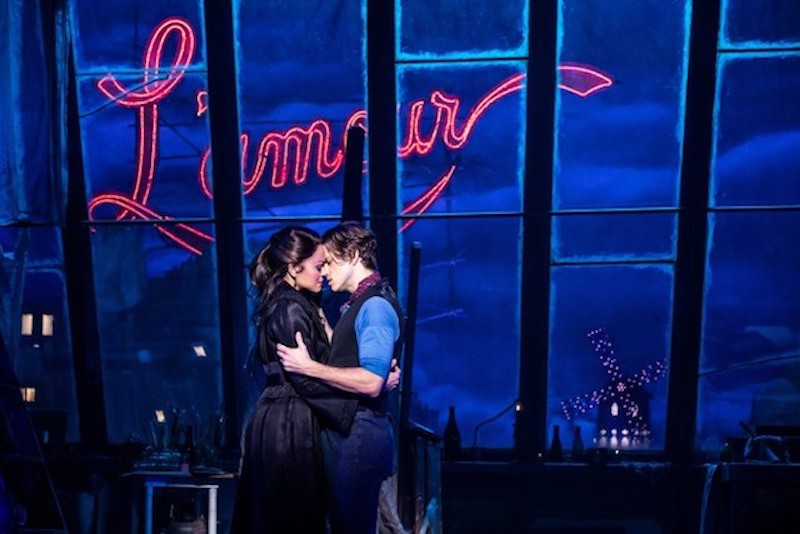 In a still form the play Moulin Rouge two actors hold each other in an embrace against windows that shine the word "L'amour" in red neon.