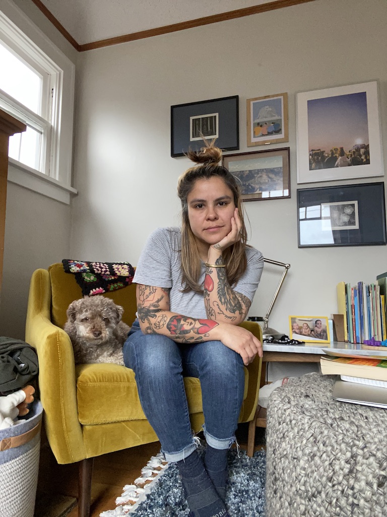 Camila and her dog sit in a yellow armchair, staring directly at the camera, her chin in her hand.