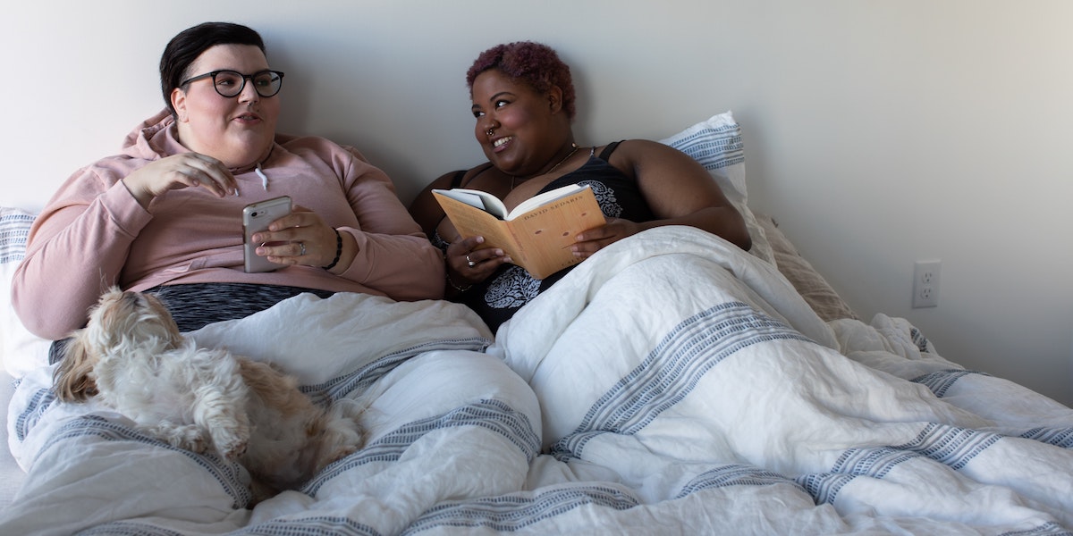 Two people curled up in bed lean into each other smiling as one reads a book and the other scrolls on their phone