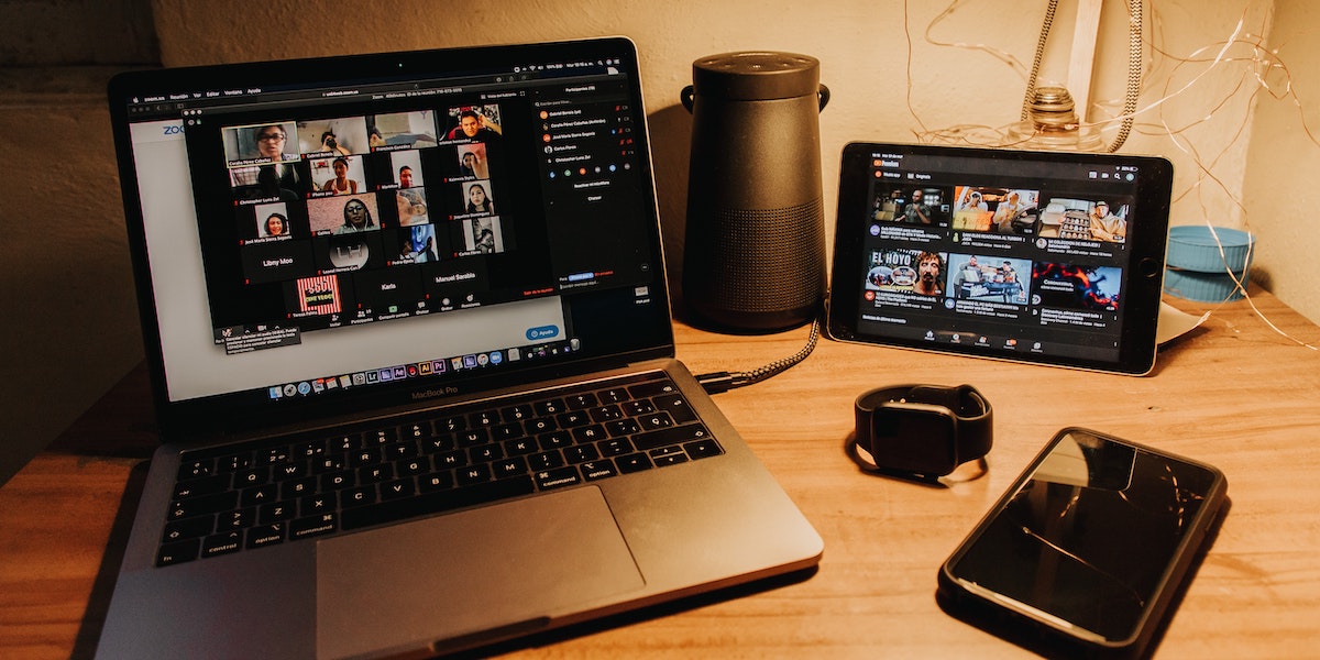 A photo of a laptop on a desk in a dark room lit by a desk lamp, with a highly populated Zoom call on the screen and a range of other electronics, a phone, apple watch and tablet, also running behind it on the same surface