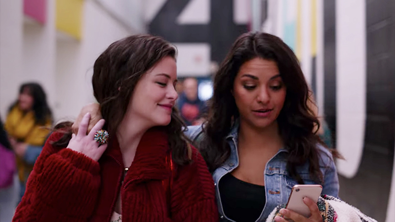 Sophie and Max from Ginny & Georgia walk down a high school hallway together, Sophie's arm is around Max's shoulder and holding Max's hand while they both look at something on Sophie's phone.