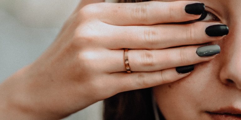 a close-up shot of a femme person with long, painted black nails using her hand to cover her eyes, just peeking through her fingers