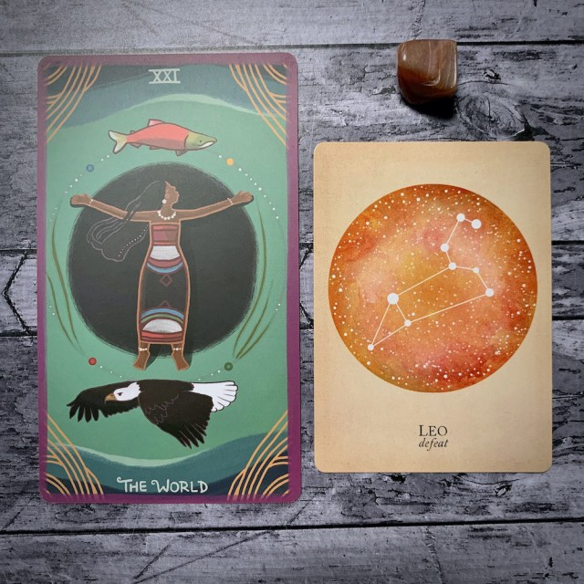 A photo of the tarot card for The World and the astrological card representing Leo, along with a small crystal