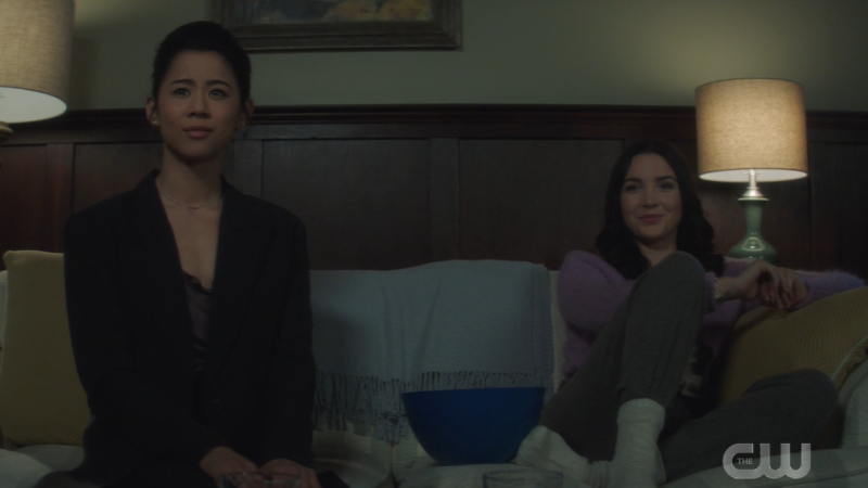 Odette in George's Body sits on the couch next to Bess, both of them purposefully not looking at each other while smiling coyly.