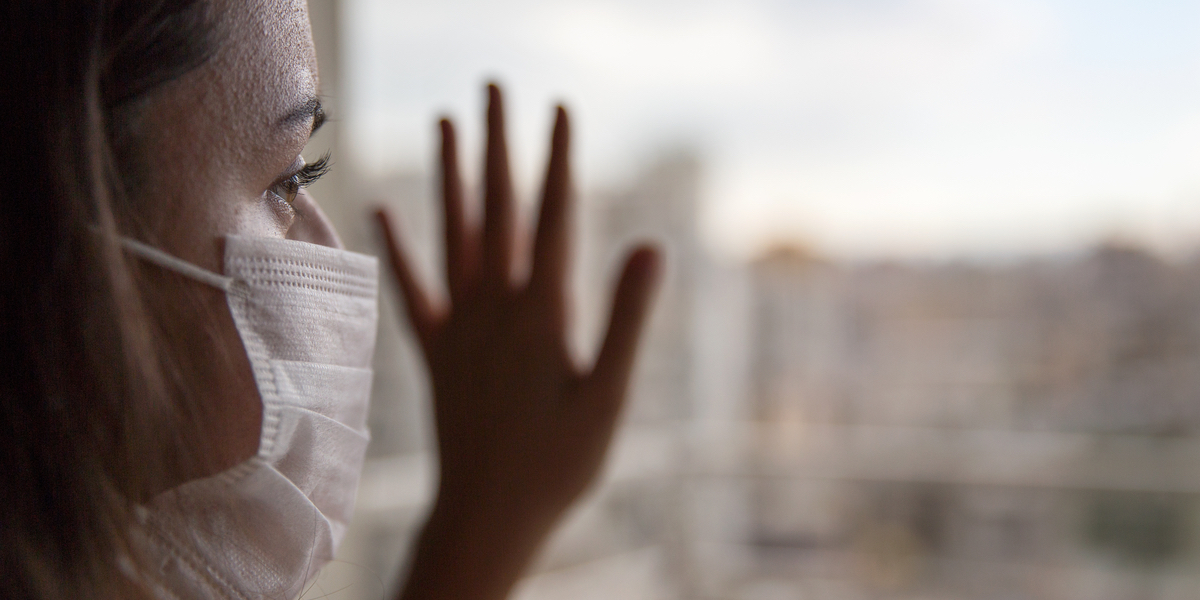 A woman with long hair wearing a medical mask presses her hand to a window and looks outside