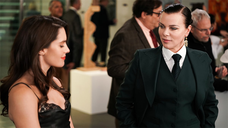 Debi Mazar as Maggie on Younger in a three-piece black suit talking to a woman in a cocktail dress at a party. 