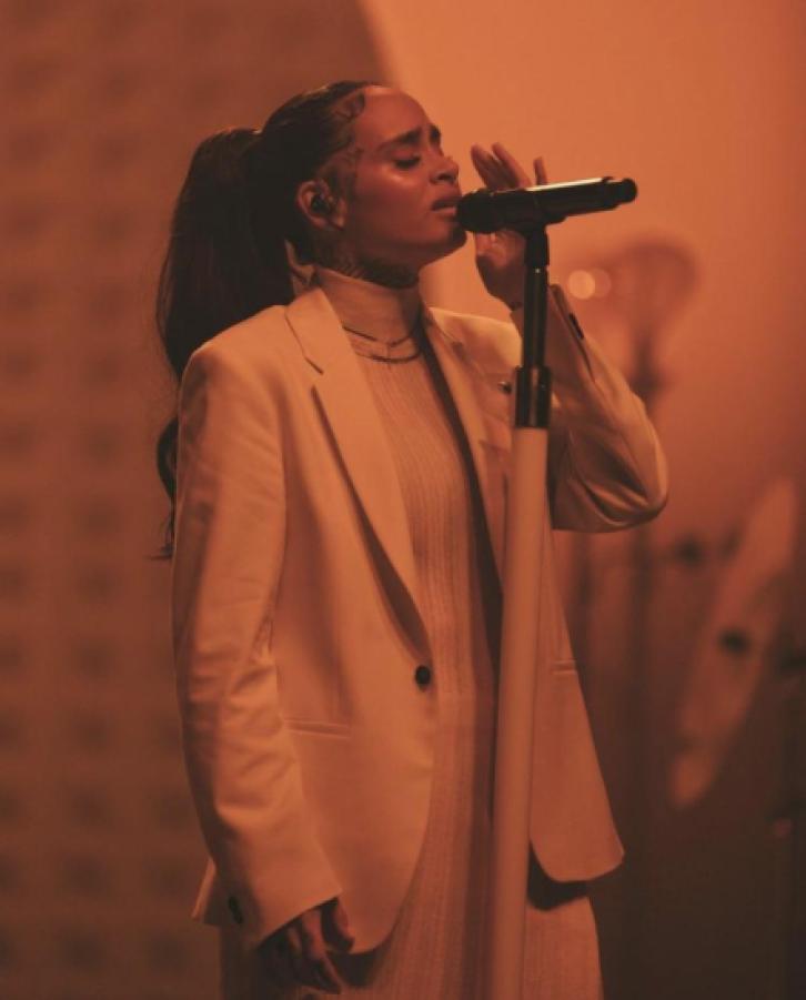 Image shows Kehlani in an all white suit holding a microphone, the lighting is red surrounding them.