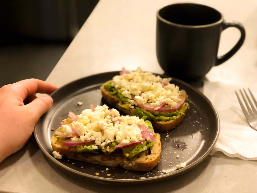 A closeup photo of two slices of avocado toast on a gray plate with a matching gray coffee cup, which Rachel's hand steadies on a kitchen counter