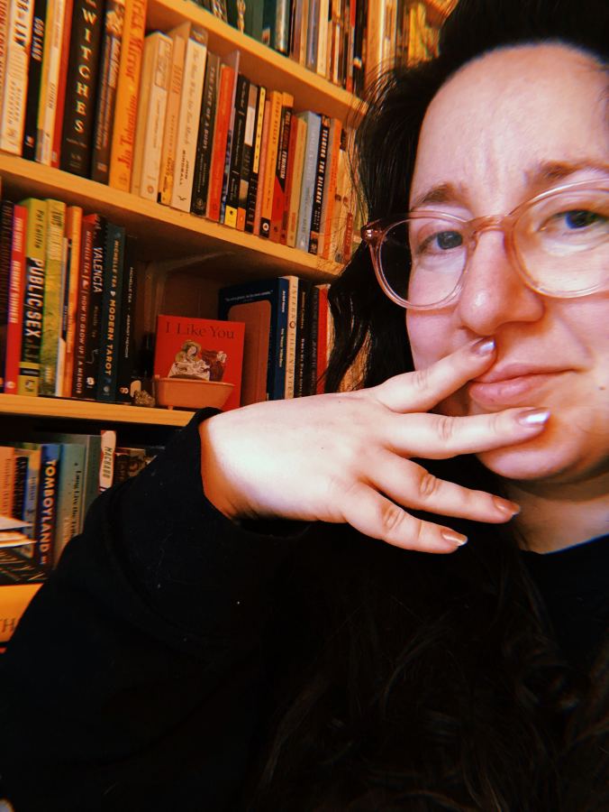 Vanessa faces the camera, hand over mouth, wearing a black shirt. Vanessa is a white woman, with glasses and long brown hair.