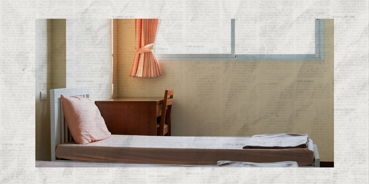 A photo of a sparse dorm-style twin bed next to a window with a wooden dorm side desk and table