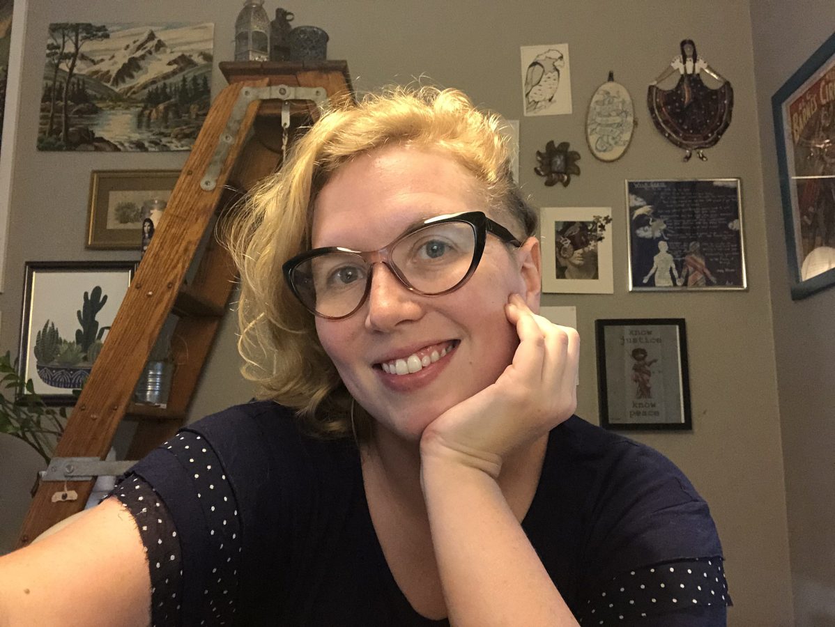 Person smiling in foreground with a blonde asymmetrical haircut and glasses, ladder and wall-mounted framed prints behind them