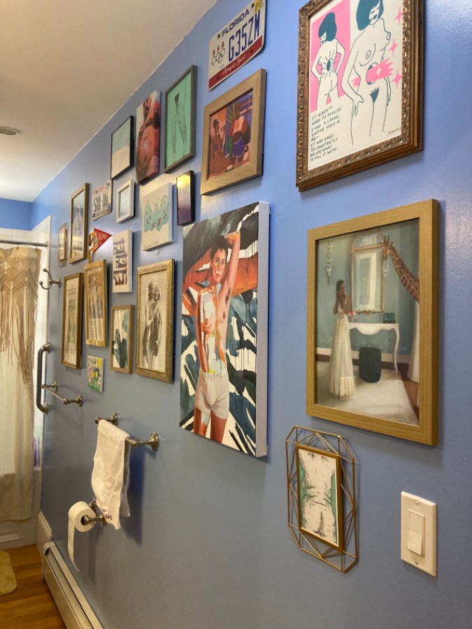 Powder blue bathroom wall covered in a variety of paintings and prints, including the figure with their hand in boxer shorts reading HANDSOME on the waistband