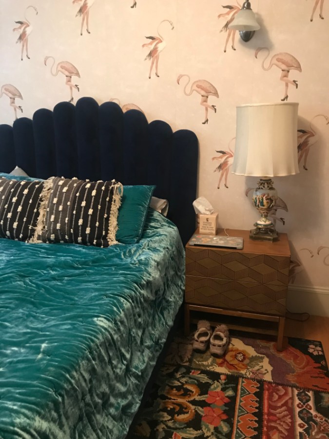 Bedroom with pale pink wallpaper covered in flamingos with human legs wearing black high heels, a dark blue headboard, a deep turquoise velvet bedspread, colorful rug, and nightstand with a lamp