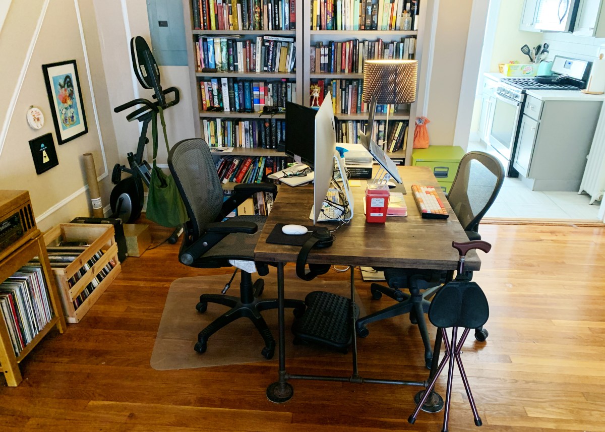 Dining table set up as a desk with a cane, stool, and medical supplies visible in addition to computer screen, bookshelves, records, and desk chair.