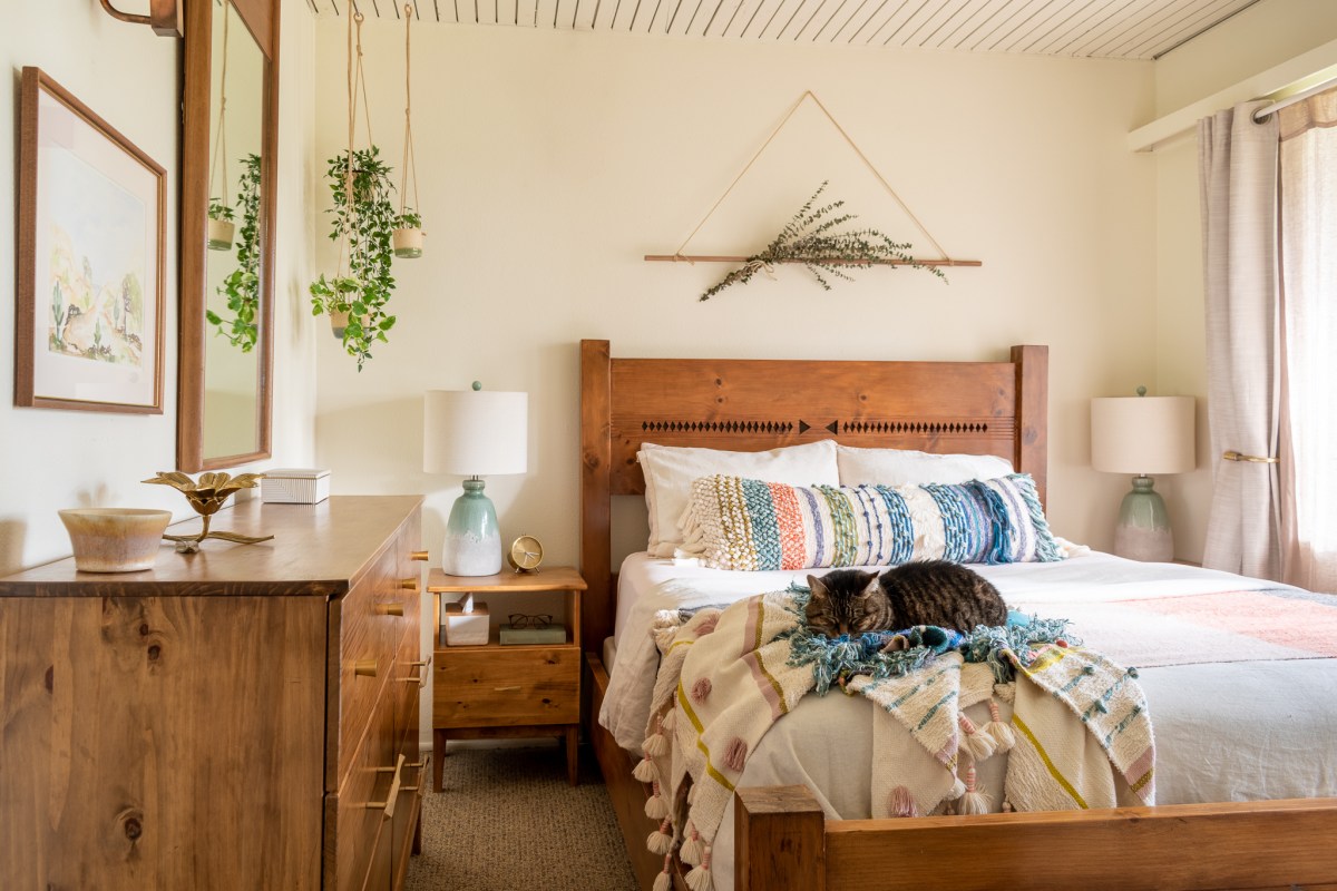 A warm, light bedroom with pale beige walls, a medium-toned bed frame covered with white bedding and colorful striped pillows, a dresser and mirror on the left side of the frame, and plants hanging on the walls. A striped cat is curled up on the blankets