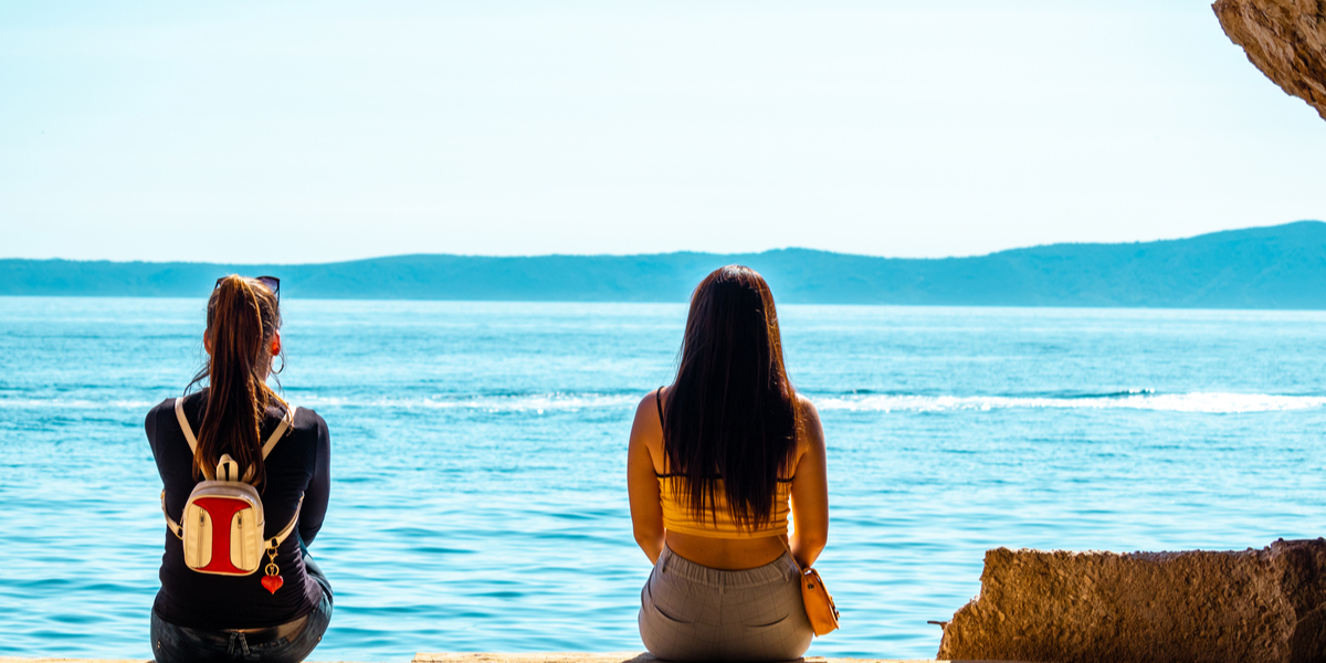 Two women sit far apart together on a beach