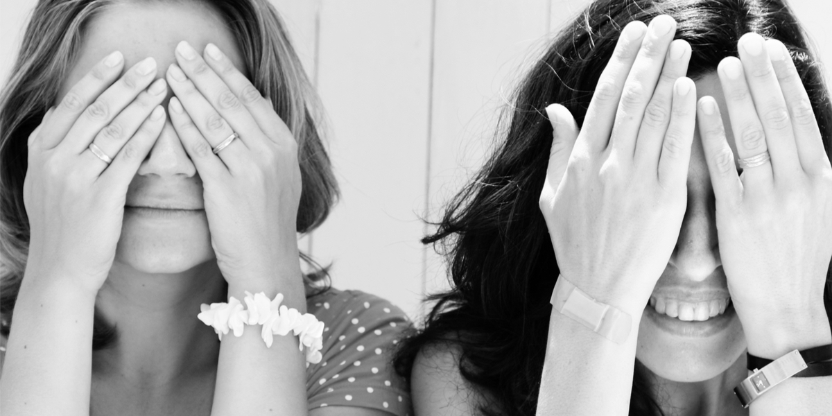 A black-and-white photo of two teenage best friends, one Latinx and one white, with their hands over their eyes and smiling at the camera
