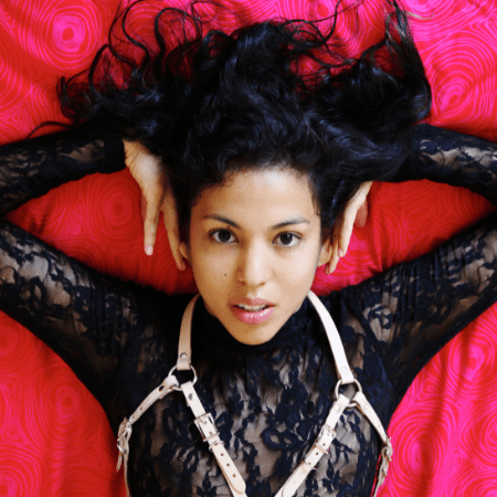 Lina Bembe is on against a red background wearing a black lace top and a white chest harness