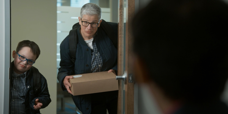 Abby stands in the doorway of an office with her nephew Mattie. She's holding a cake. They both wear glasses.