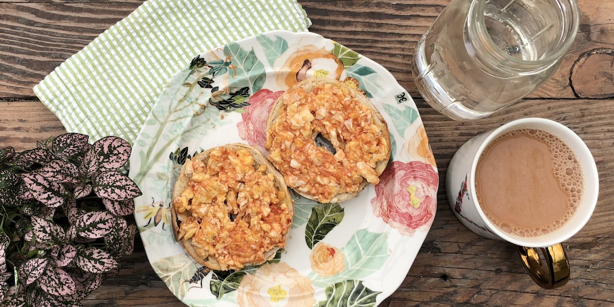 a breakfast spread including a bagel with scrambled eggs on a floral print plate, a mason jar full of water, a cup of tea, a cute plant, and a green and white striped napkin all set on a wood table