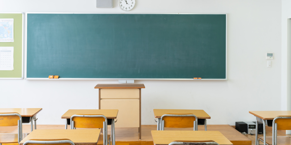 An empty classroom, which includes wooden desks, a green chalkboard, a clock and a podium at the front of the room.