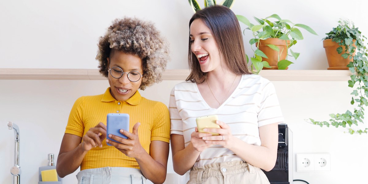 two individuals sit on a couch looking at their phones as though they are looking at an advice column. the one on the left inspects the screen. they are Black, have hair in a light brown afro style, are wearing glasses and a yellow collared shirt. next to them is a person who is looking at their screen and grinning. this person is taller, white, has long straight brown hair and is wearing a white striped tee