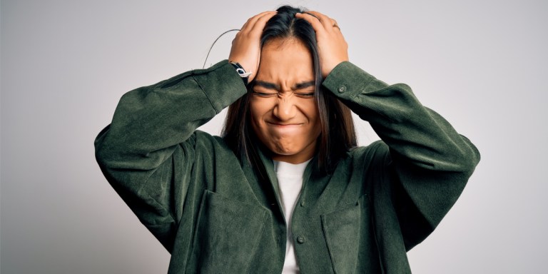 A south asian person with long her and a baggy jacket is frustrated with their hands on their head