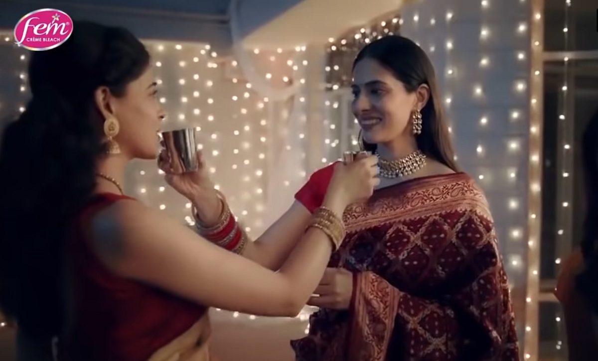 Two women are holding up silver cups of water to each other to drink from. They are both wearing saris, dressed in red and gold.