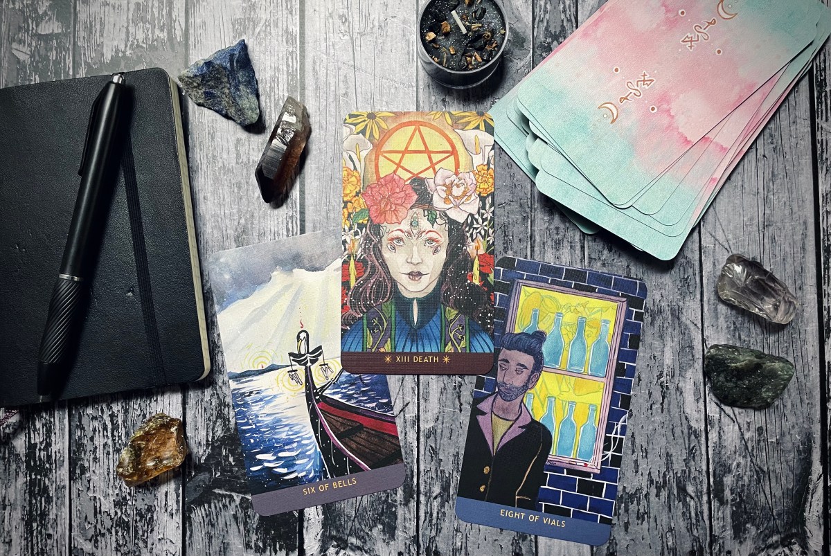 gorgeous tarot spread on a wood table, includes a notebook and pen, a tarot deck, multiple crystals, and three pulled cards: six of bells, death, and eight of vials