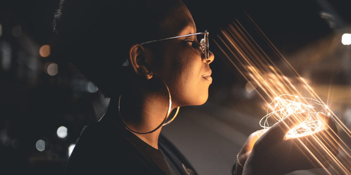 Black woman with large gold hoop earrings and natural hair closes her eyes as though she is making a wish on the christmas lights she is holding in her hand,