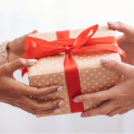 A person with long nails hands a present wrapped in brown paper with white polka dots and a red ribbon to a person with long nails that have been painted lavender.