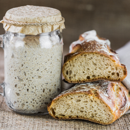 A covered mason jar containing a sourdough starter sits beside a sourdough baguette