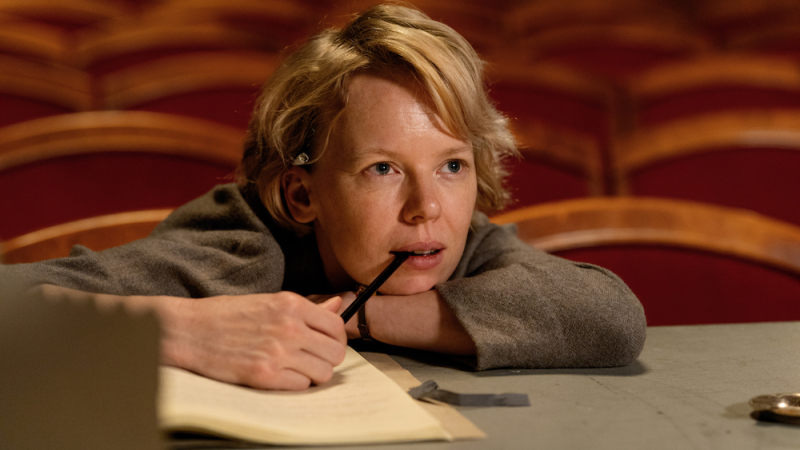 Alma Poystï looks up at a stage with a pen in her mouth, hand resting on a notebook.
