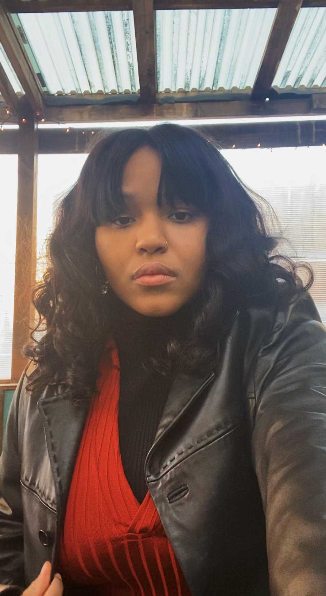 This is a photo of Amari Gaiter. Amari is a Black person, and in this photo has long, loosely curled black or dark brown hair, and is wearing a nose ring, earrings, a red sweater and a leather jacket. They are looking directly at the camera with a focused expression.