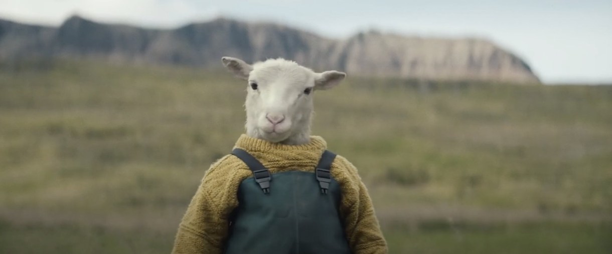 This is an image of a white lamb that appears to be standing upright although we can only see it from the waist up. It is wearing a yellow chunky knit sweater and gray coveralls. There are mountains in the background.