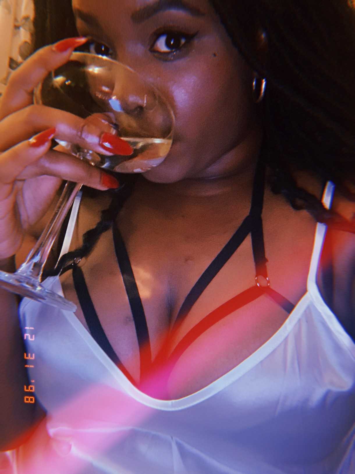 Shelli is drinking a cocktail. Shelli is wearing a strappy decorative bra under a white tank top and has long red nails.