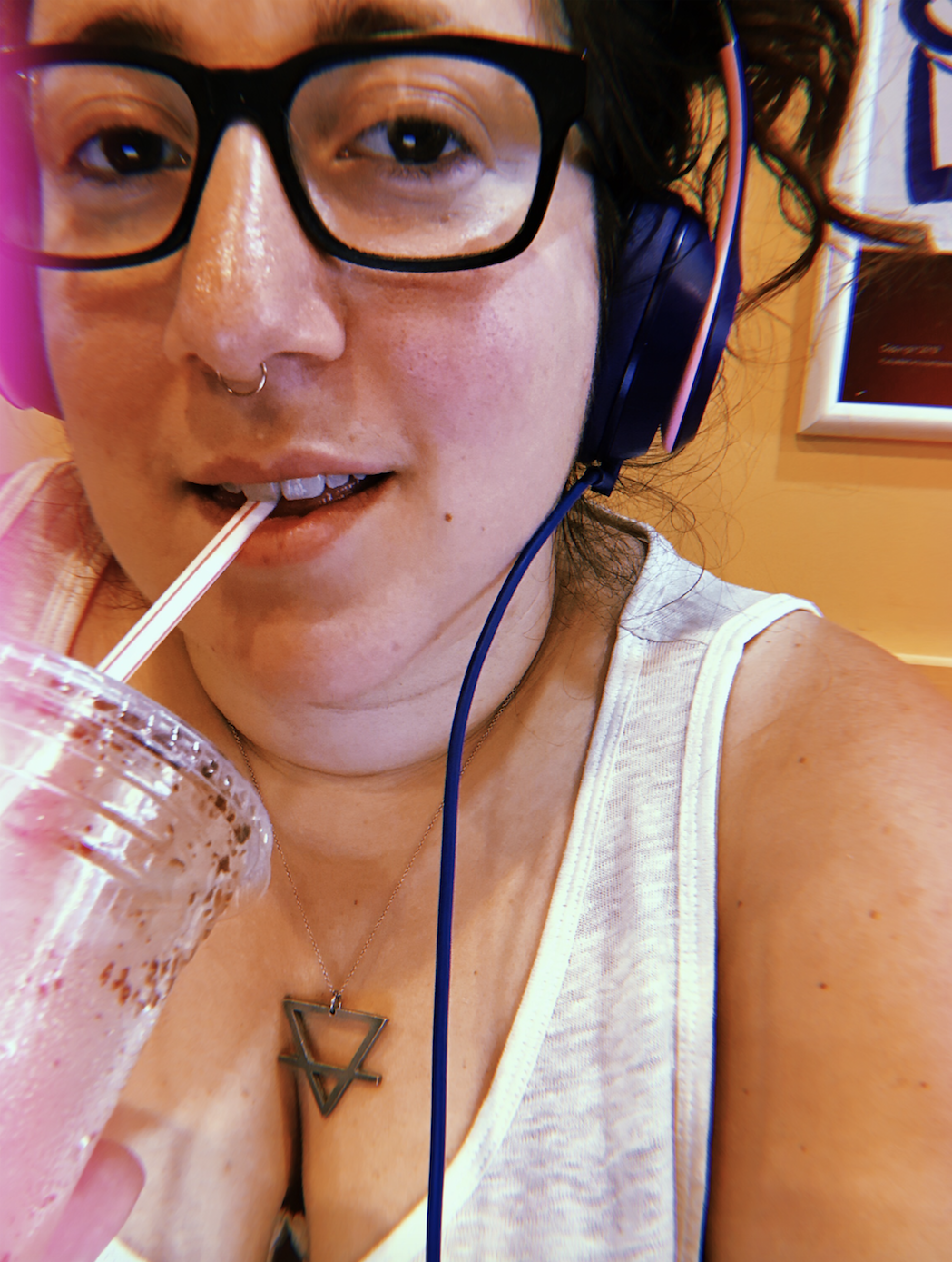 Vanessa is drinking an iced coffee and wearing an earth symbol necklace. She wears glasses.