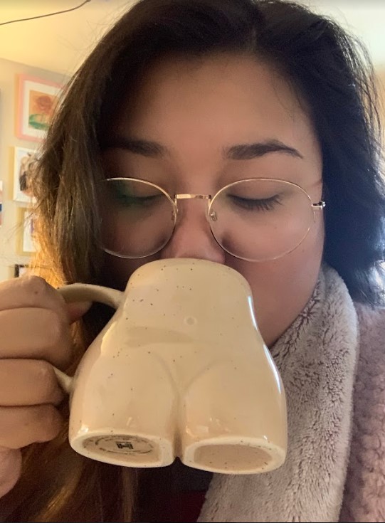 Writer, Em, drinks bourbon maple coffee out of a mug that is shaped like the lower torso of a person. Em has long brown hair and glasses.