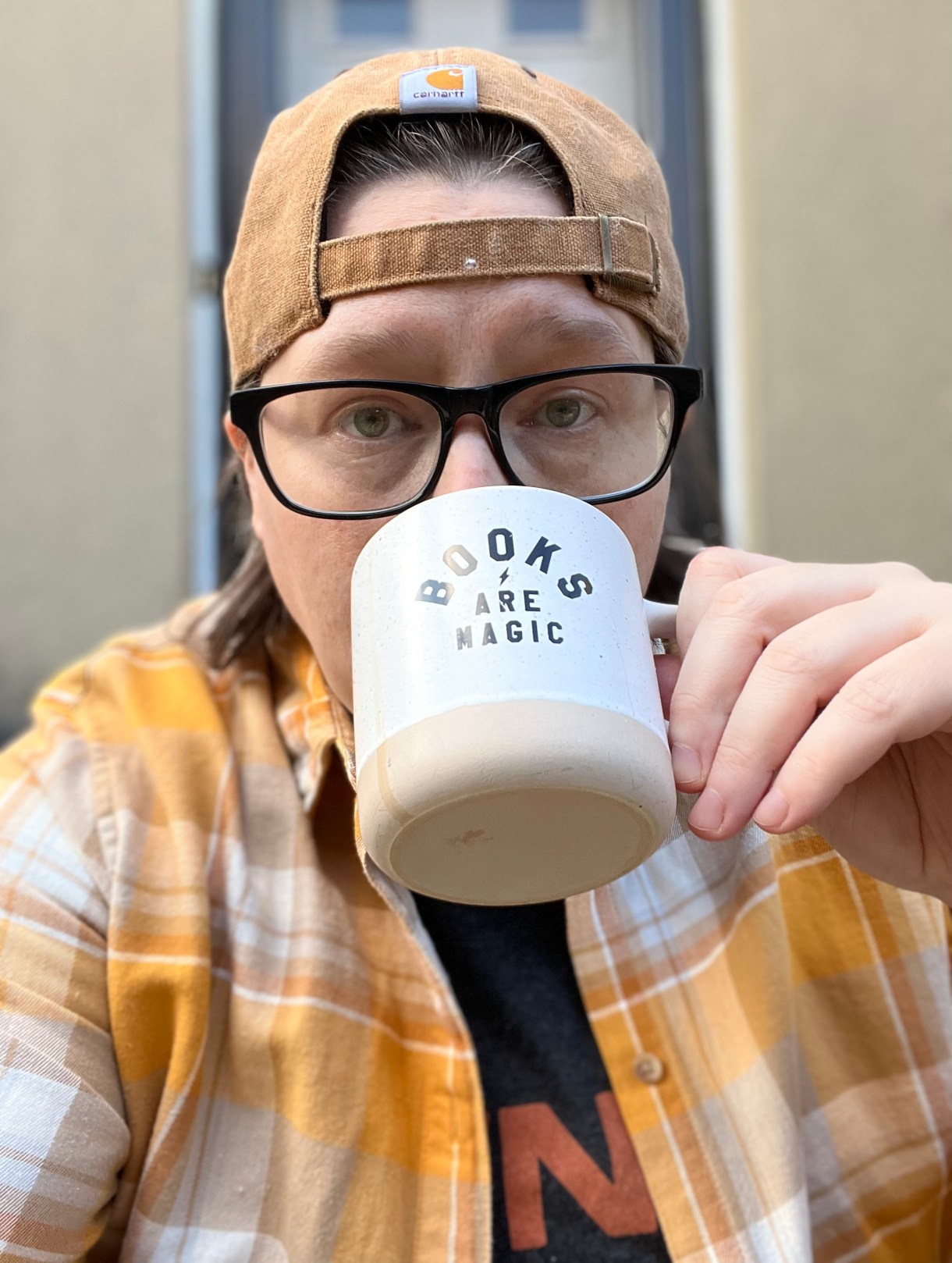 Heather drinking out of a Books Are Magic mug. Heather is wearing a backwards baseball cap. Heather wears glasses.