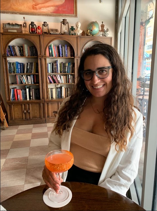 Kayla is drinking a cocktail and smiling in a room that looks like it may be in a bookstore. The cocktail looks fruity. Kayla has long wavy brown hair and glasses.