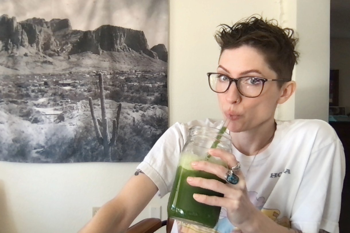 Laneia is drinking a green juice. Laneia has short brown hair and is wearing a white tee. Laneia wears glasses.