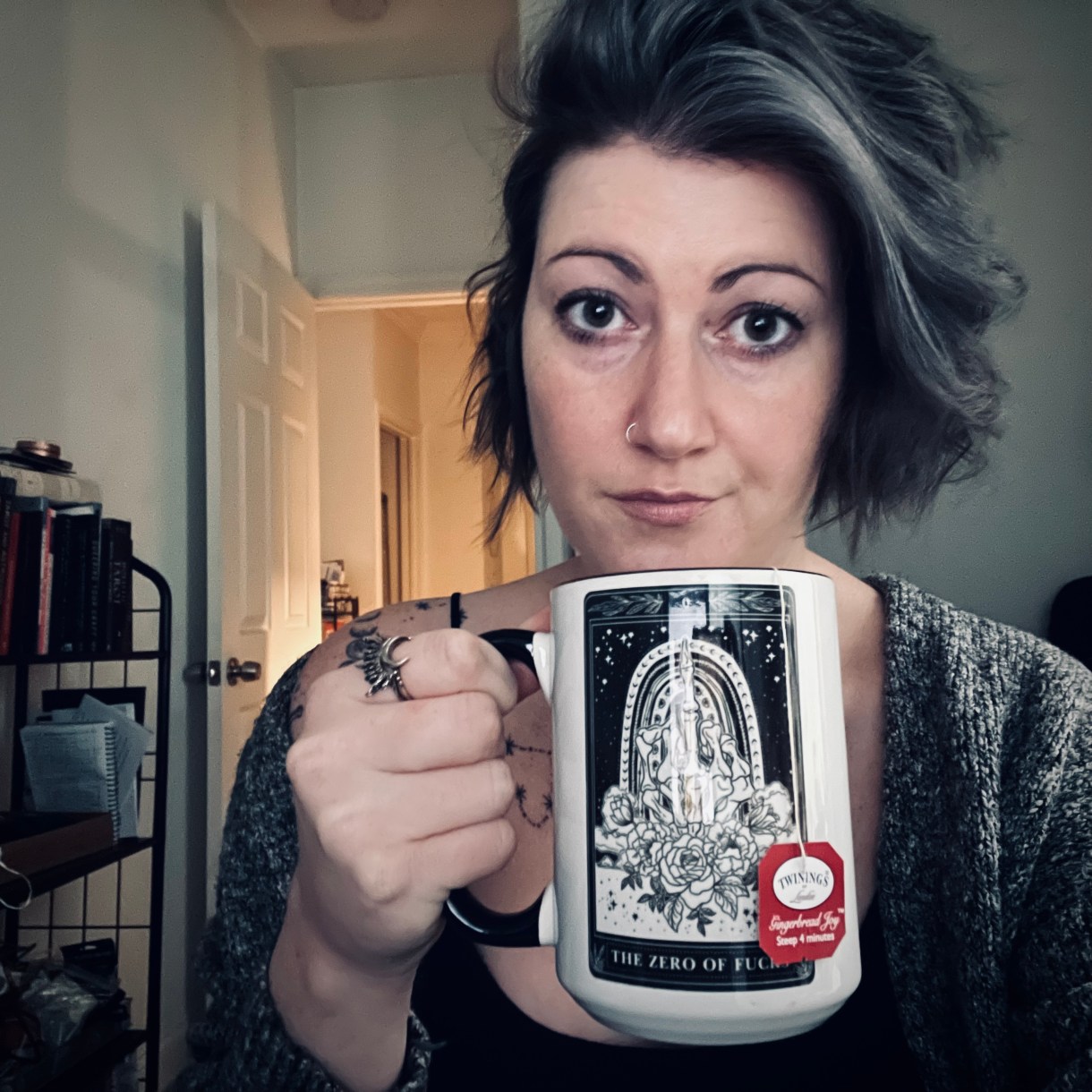 Meg is drinking out of a tarot themed mug. Mug has light skin and black hair streaked with gray. Meg does NOT wear glasses.