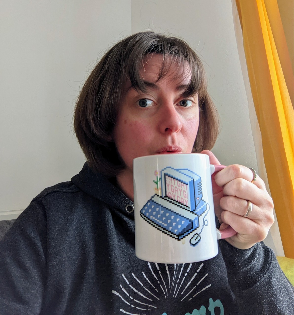 Sally is drinking out of a Gay Computer Autostraddle fundraiser mug. Sally has bangs and a bob-style haircut. Her hair is brown.