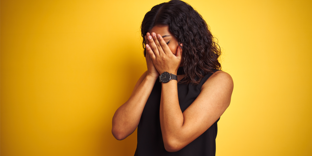 Against a yellow background, a woman with brown skin and shoulder-length, wavy brown hair puts her hands over her face. She is wearing a sleeveless black shirt and a black watch.