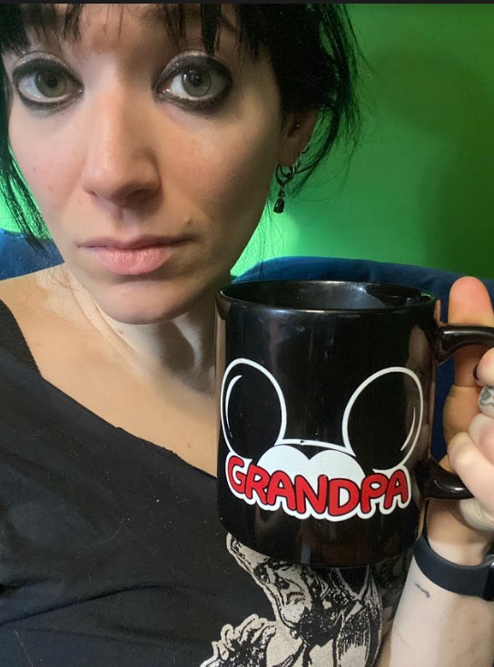 Stef is drinking hot cocoa out of a disney mug that says Grandpa. Stef is wearing a shirt with a Frankenstein's monster on it.