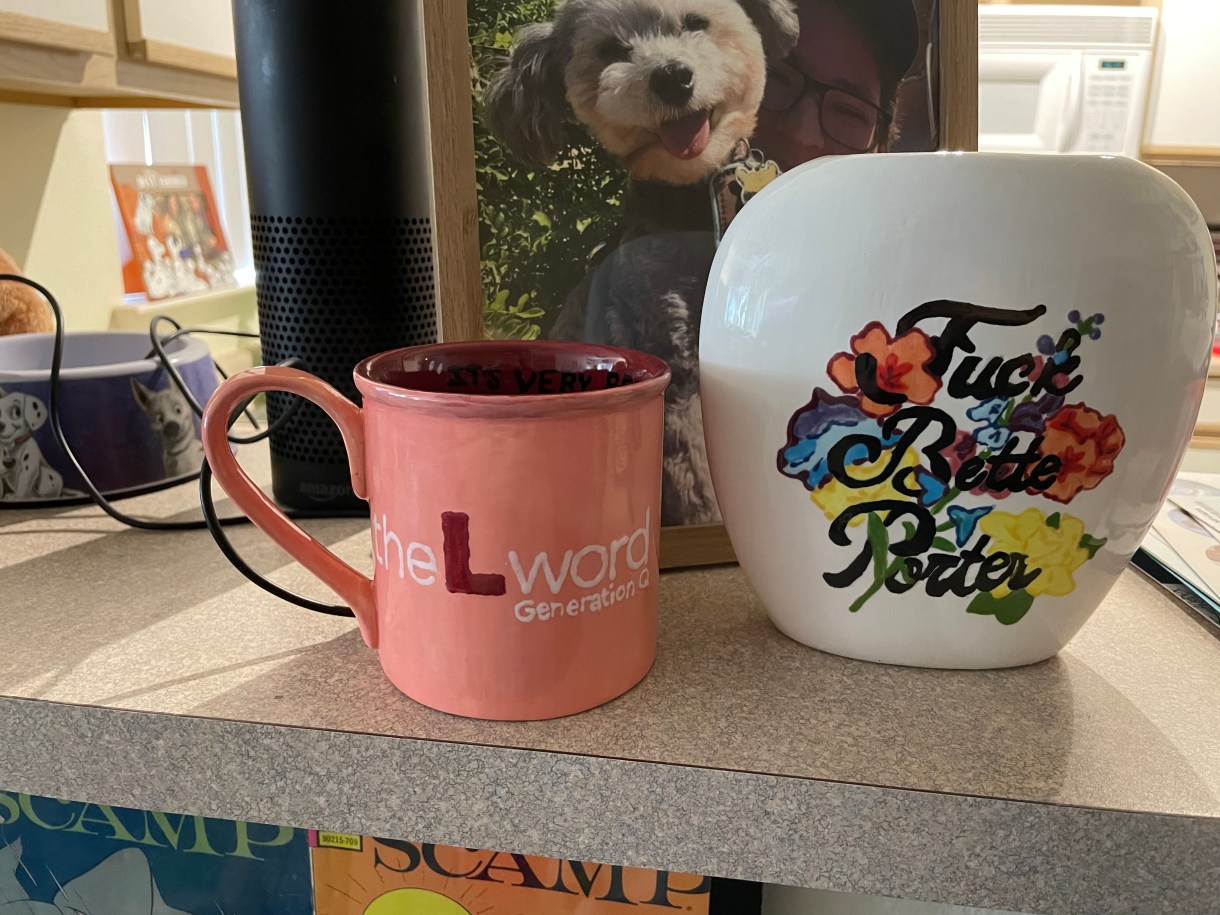 This is the second view of two pieces of pottery. On the left is a mug the same mug, reading on the other side "The L Word Generation Q" and which has a heart on it. On the right is a vase that says "Fuck Bette Porter" over a floral background.