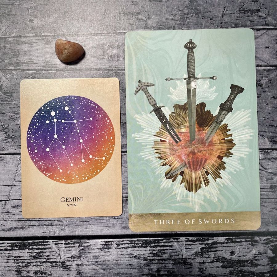 A card says gemini: unite with an purple circle, next to a card that says three of swords with an illustration of three swords stabbing a heart