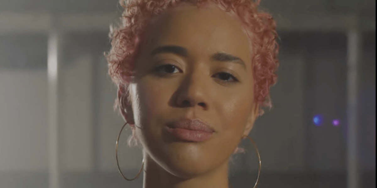 A close up of Jasmin, wearing a white shirt, large hoop earrings and her pink tinged hair is also in the frame.