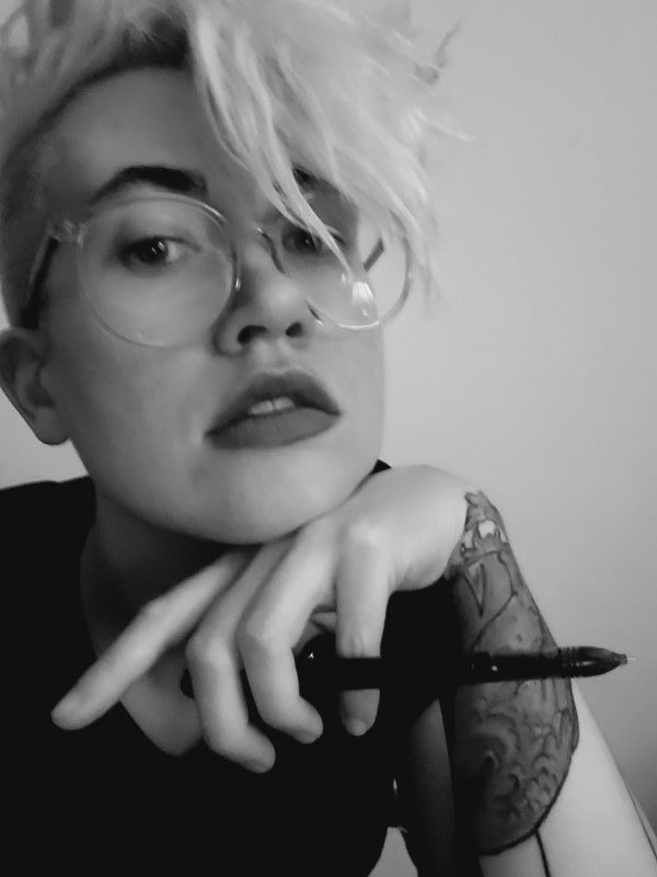 Here's Nicole leaning on their hand, which is holding a pen. They are a white, genderqueer human with bleached hair, shaved sides of the hair, large clear glasses, a tattoo sleeve and dark eyebrows. The photo is in black and white.