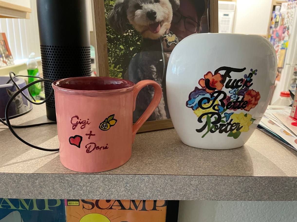 This is the first view of two pieces of pottery. On the left is a mug that says "Gigi + Dani" and which has a heart on it. On the right is a vase that says "Fuck Bette Porter" over a floral background.